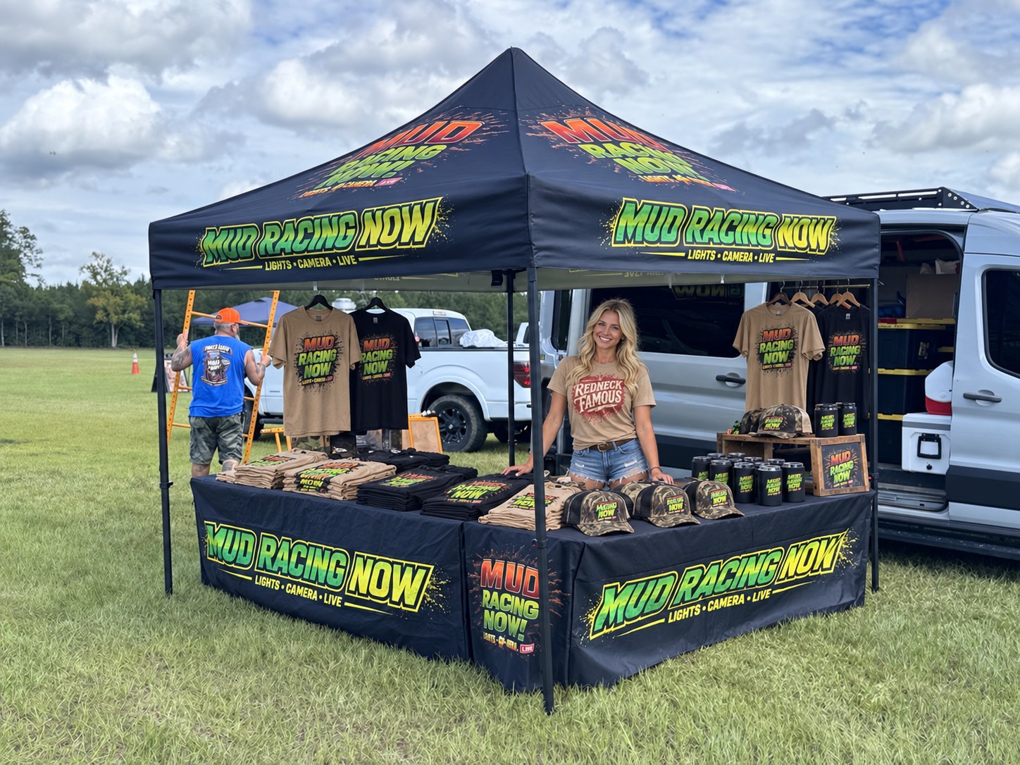 Stocked Mud Racing NOW merchandise tent with shirts, hats, drinkware, and an adult saleswoman wearing Redneck Famous apparel